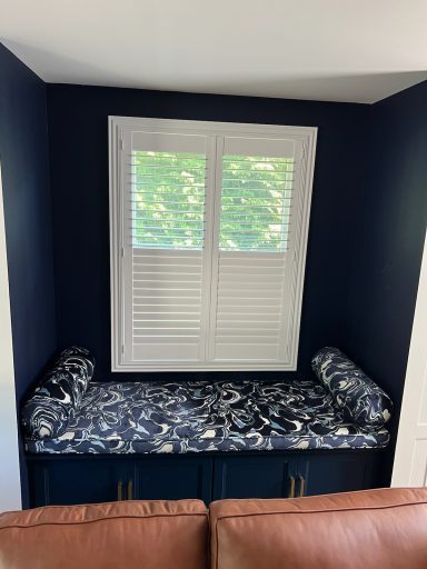 Cozy window nook with a patterned cushioned bench and white shutters.